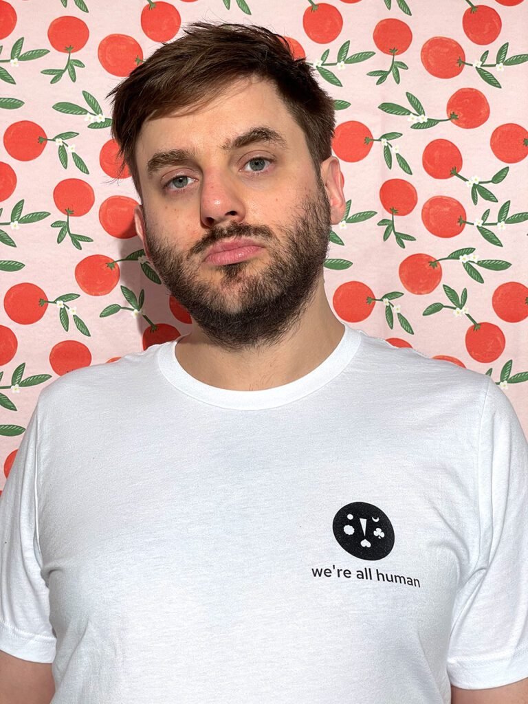 Mark, a white-skinned man with short brown wavy hair and grey eyes. He’s looking at the camera and wearing a white t-shirt with the we’re all human logo on. 