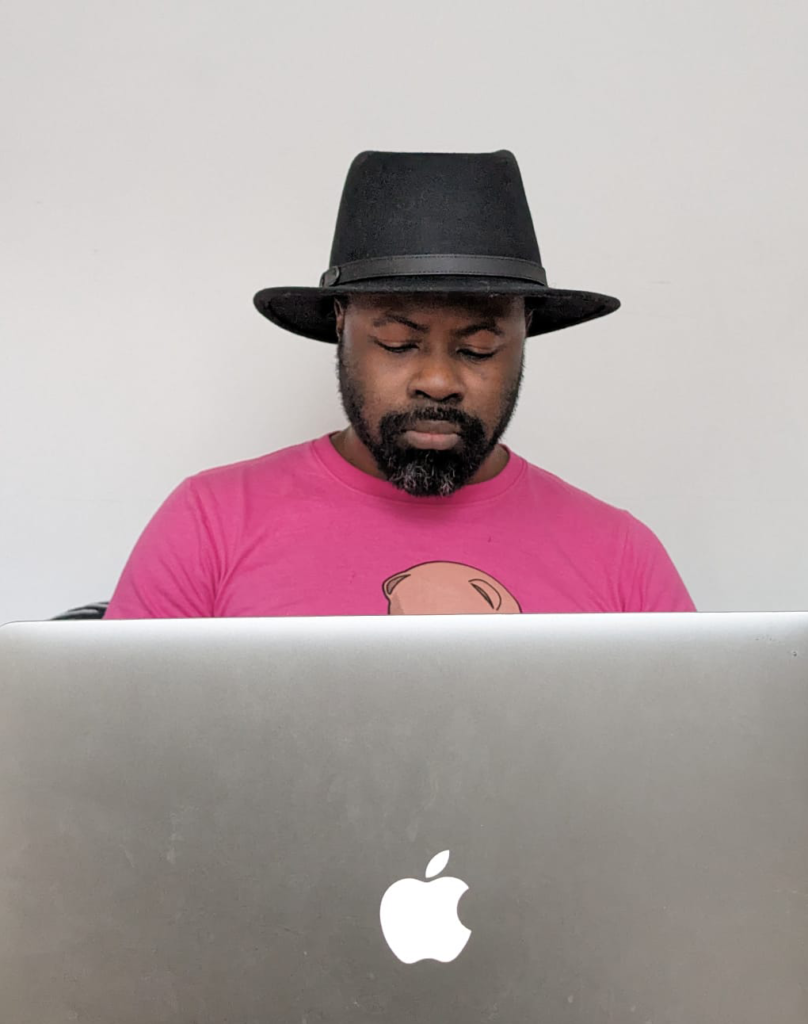 A photo of myself siting in front of a laptop with a pink t-shirt and a black fedora hat