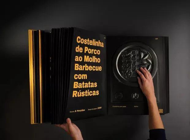 A large book with bold yellow type on black pages. A hand reaches out to touch the second page, embossed with the shape of a plate with foodstuff